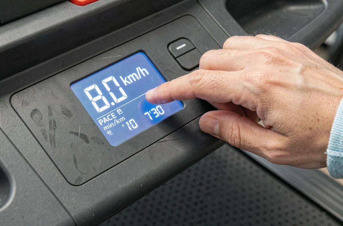 Gros plan sur la console d'un tapis de course affichant 8 km/h et la conversion en min/km, main ajustant la vitesse